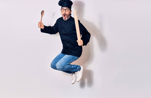 Middle age handsome hispanic man surprised wearing glasses and cook uniform. Holding kneader and spoon jumping over isolated white background - Foto, immagini