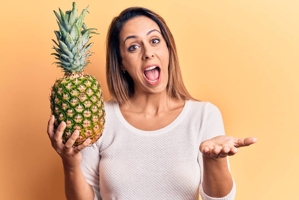 Young beautiful woman holding pineapple celebrating achievement with happy smile and winner expression with raised hand  - Photo, Image