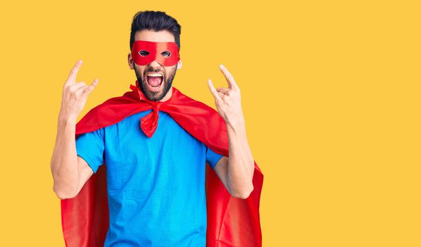 Young handsome man with beard wearing super hero costume shouting with crazy expression doing rock symbol with hands up. music star. heavy concept.  - Photo, Image