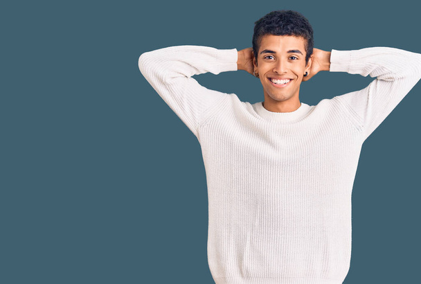 Jeune homme afro-américain portant des vêtements décontractés relaxant et étirant, les bras et les mains derrière la tête et le cou souriant heureux  - Photo, image