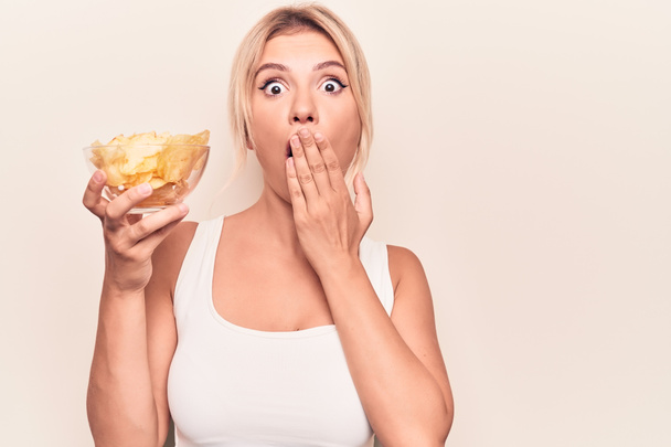 Junge schöne blonde Frau hält Schüssel mit Kartoffelchips über isolierten weißen Hintergrund, der Mund mit der Hand bedeckt, schockiert und Angst vor Fehler. Überraschte Miene - Foto, Bild