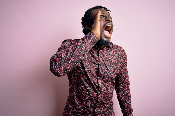 Jovem homem afro-americano bonito vestindo camisa casual em pé sobre fundo rosa gritando e gritando alto para o lado com a mão na boca. Conceito de comunicação
. - Foto, Imagem
