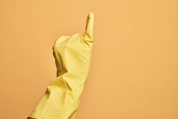 Mano del joven caucásico con guante de limpieza sobre fondo amarillo aislado mostrando el dedo meñique como compromiso de promesa meñique, número uno - Foto, imagen