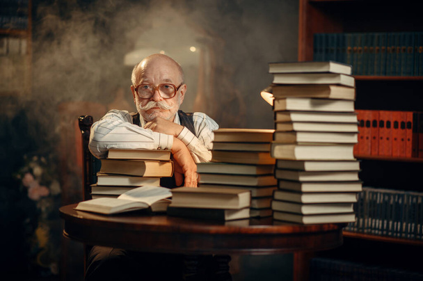 Escritor de edad avanzada sentado en la mesa con un montón de libros en la oficina del hogar. Viejo con gafas escribe novela literaria en la habitación con humo, inspiración - Foto, Imagen