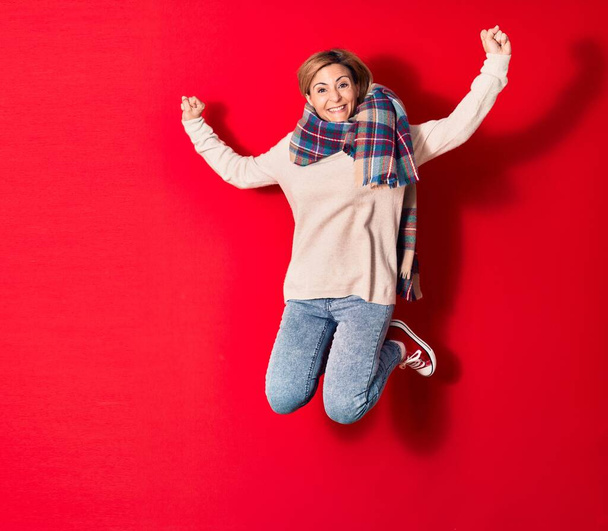 Νεαρή όμορφη γυναίκα που φοράει καθημερινά χειμωνιάτικα ρούχα χαμογελώντας ευτυχισμένη. Άλμα με χαμόγελο στο πρόσωπο πάνω από απομονωμένο κόκκινο φόντο - Φωτογραφία, εικόνα