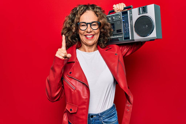 Mujer hipster morena de mediana edad sosteniendo música retro boombox sobre fondo rojo aislado sonriendo con una idea o pregunta señalando el dedo con la cara feliz, número uno
 - Foto, Imagen