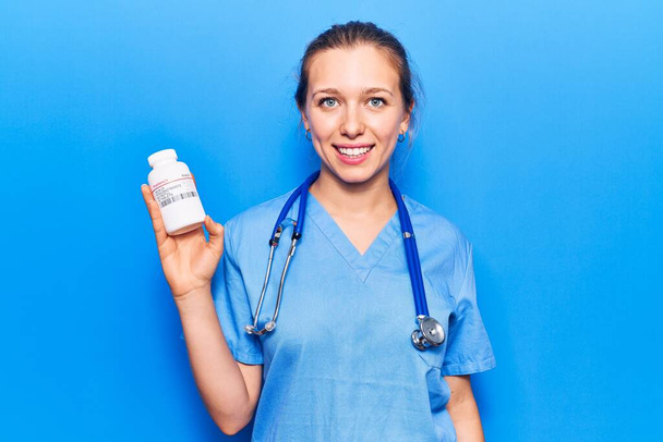 Молода красива блондинка в уніформі лікаря тримає таблетки, виглядає позитивно і щасливо стоячи і посміхаючись з впевненою посмішкою показуючи зуби
  - Фото, зображення