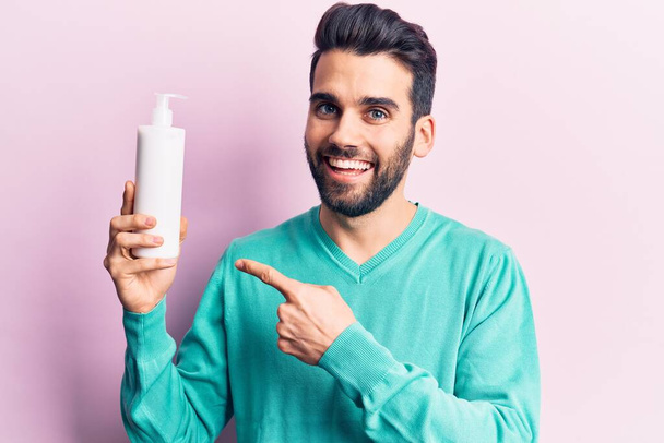 Young handsome man with beard holding bottle of cream smiling happy pointing with hand and finger  - Fotó, kép