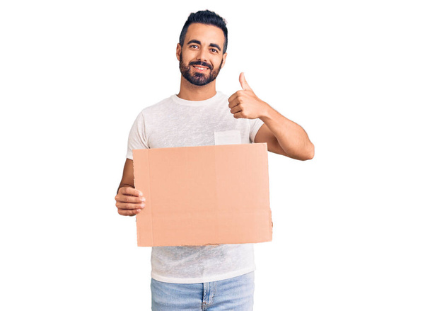 Young hispanic man holding banner cardboard smiling happy and positive, thumb up doing excellent and approval sign  - Φωτογραφία, εικόνα