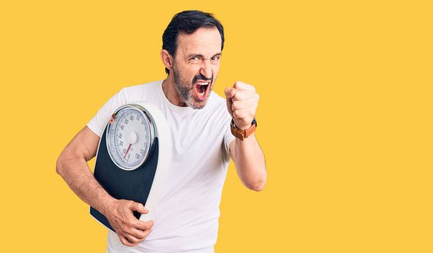 Middle age handsome man holding weighing machine annoyed and frustrated shouting with anger, yelling crazy with anger and hand raised  - Фото, изображение