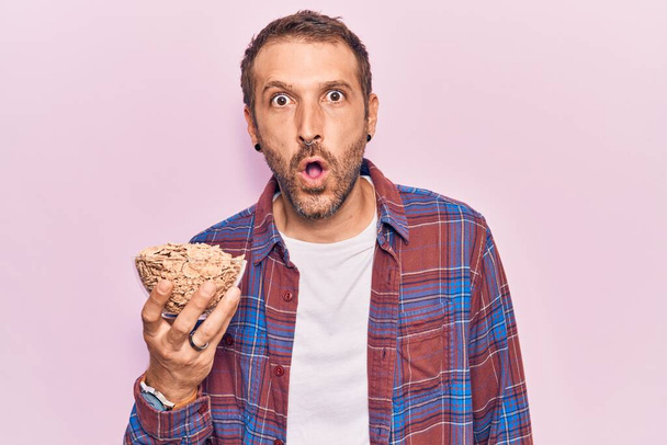 Young handsome man holding cornflakes scared and amazed with open mouth for surprise, disbelief face  - Photo, Image