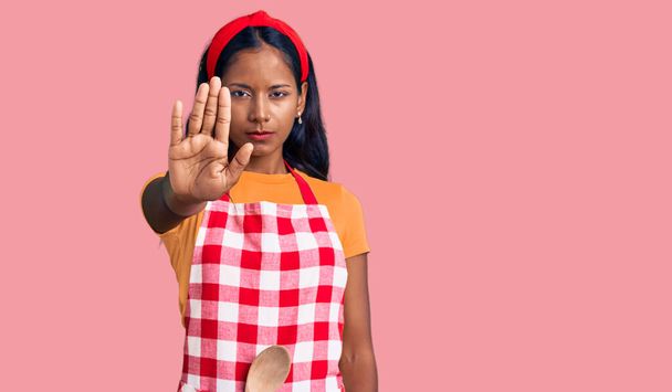 Young indian girl wearing professional baker apron doing stop sing with palm of the hand. warning expression with negative and serious gesture on the face.  - Photo, Image