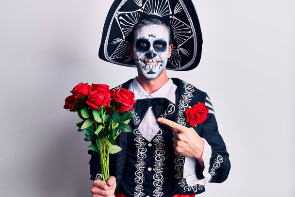 Young man wearing mexican day of the dead costume holding flowers smiling happy pointing with hand and finger  - Photo, Image