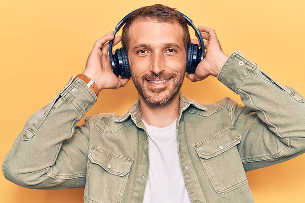 Молодий красивий чоловік слухає музику за допомогою навушників виглядає позитивним і щасливим стоячи і посміхаючись з впевненою посмішкою, показуючи зуби
  - Фото, зображення
