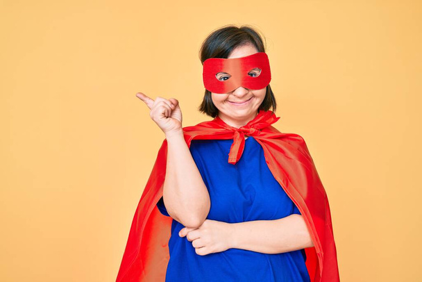 Donna bruna con la sindrome di Down indossando costume da super eroe sorridente felice indicando con mano e dito di lato  - Foto, immagini