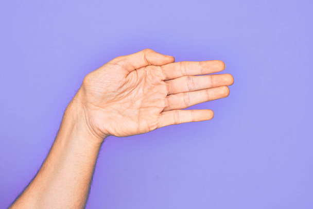 Ręka białego młodzieńca pokazuje palce nad odizolowanym fioletowym tłem rozciągającym się i sięgającym z otwartą ręką po uścisk dłoni, pokazując dłoń - Zdjęcie, obraz
