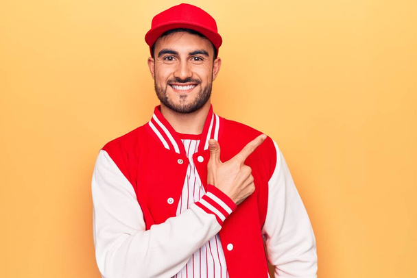 Hombre deportivo guapo con barba usando chaqueta de béisbol y gorra sobre fondo amarillo sonriendo alegre señalando con la mano y el dedo hacia el lado
 - Foto, imagen