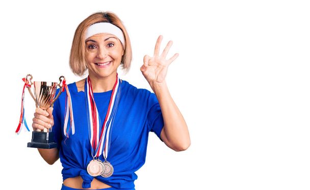 Fiatal szőke nő kezében bajnok trófea visel medál csinál rendben jel ujjakkal, mosolygós barátságos gesztus kiváló szimbólum  - Fotó, kép