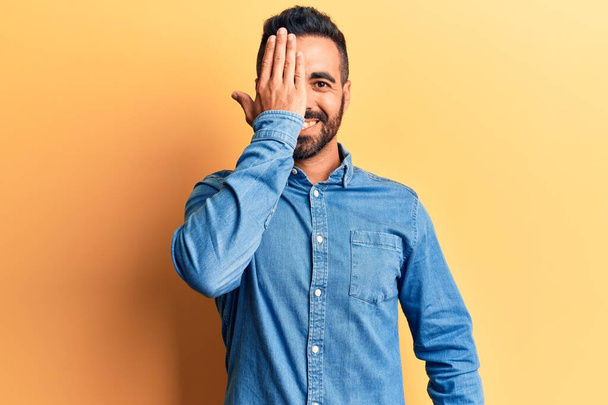 Young hispanic man wearing casual clothes covering one eye with hand, confident smile on face and surprise emotion.  - Photo, Image