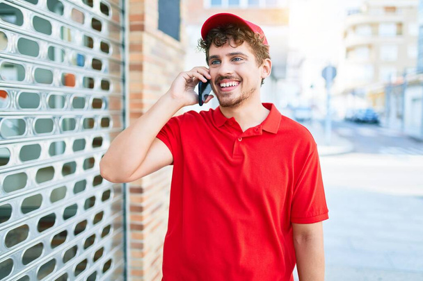 Young caucasian delivery man smiling happy having conversation talking on the smartphone at city. - 写真・画像