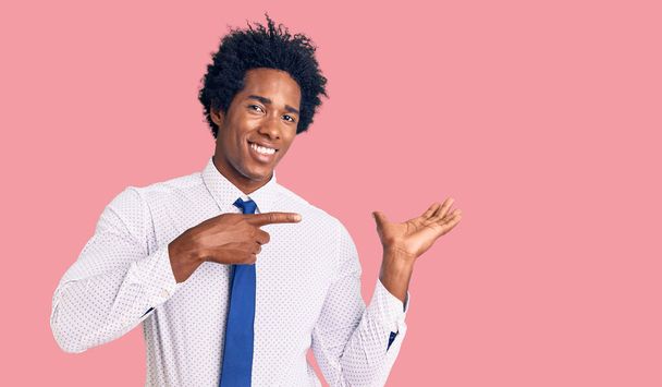 Bonito homem americano africano com cabelo afro vestindo roupas de negócios maravilhado e sorrindo para a câmera enquanto se apresenta com a mão e apontando com o dedo
.  - Foto, Imagem