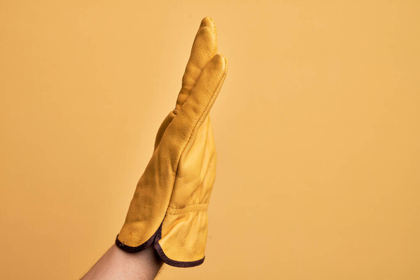 Bahçıvan eldiveni takmış beyaz bir gencin eli izole edilmiş sarı arka planda uzanmış el hareketlerini gösteriyor. - Fotoğraf, Görsel