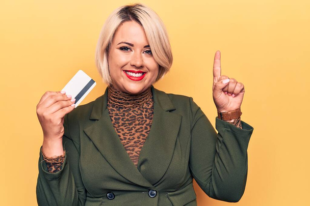 Jovem linda loira plus size mulher segurando cartão de crédito sobre fundo amarelo isolado sorrindo com uma ideia ou pergunta apontando dedo com rosto feliz, número um
 - Foto, Imagem