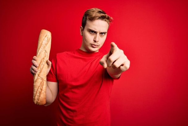 Jovem homem ruivo bonito segurando pão caseiro fresco sobre fundo vermelho isolado apontando com o dedo para a câmera e para você, gesto confiante olhando sério
 - Foto, Imagem