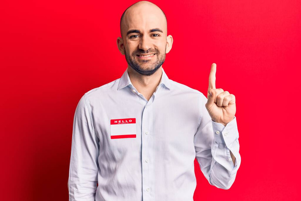 Young handsome bald man wearing hello my name is sticker smiling with an idea or question pointing finger with happy face, number one  - Φωτογραφία, εικόνα