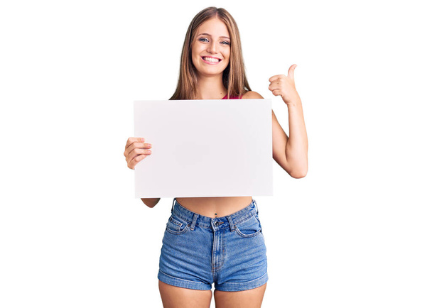 Jovem bela mulher loira segurando banner vazio em branco sorrindo feliz e positivo, polegar para cima fazendo excelente e sinal de aprovação  - Foto, Imagem