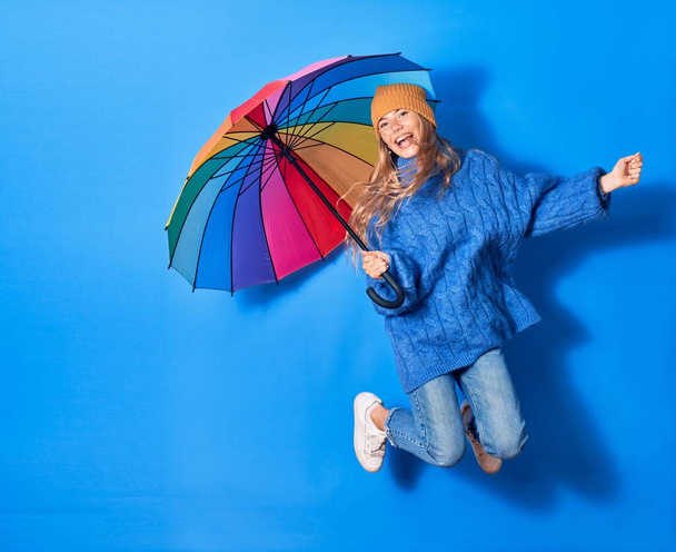 Młoda piękna kobieta w zimowych ciuchach uśmiechnięta szczęśliwa. Skoki z uśmiechem na twarzy trzymając kolorowy parasol na odizolowanym niebieskim tle - Zdjęcie, obraz