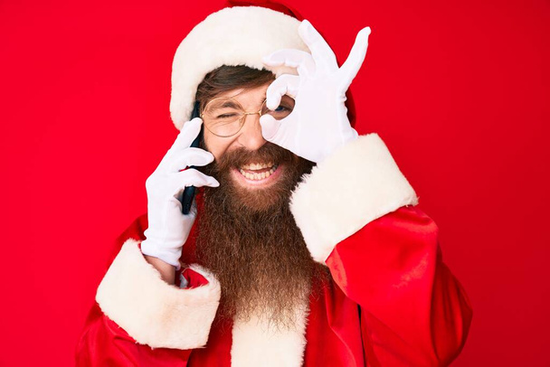 Guapo joven cabeza roja hombre con barba larga con traje de Santa Claus hablando por teléfono sonriendo feliz haciendo signo de ok con la mano en el ojo mirando a través de los dedos
  - Foto, Imagen