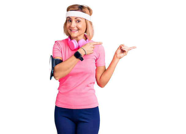 Mujer rubia joven con ropa deportiva y auriculares sonriendo y mirando a la cámara apuntando con dos manos y dedos hacia un lado
.  - Foto, Imagen
