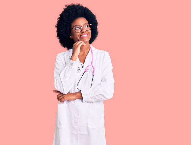 Молодая африканская американка в медицинском пальто и стетоскопе с рукой на подбородке думает о вопросе, задумчивом выражении лица. улыбающееся и вдумчивое лицо. Концепция сомнений.  - Фото, изображение