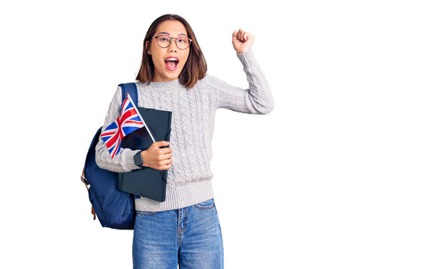 Mladý krásný číňan dívka na sobě student batoh drží pořadač a uk vlajka křičí hrdě, slaví vítězství a úspěch velmi vzrušený se zdviženými pažemi  - Fotografie, Obrázek