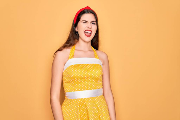 Jeune belle pin up femme portant des années 50 robe vintage de mode sur fond jaune clin d'oeil en regardant la caméra avec une expression sexy, gai et heureux visage. - Photo, image