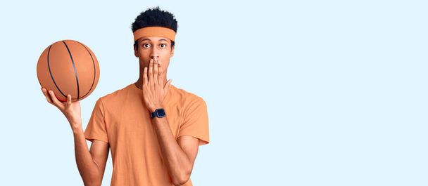 Young african american man holding basketball ball covering mouth with hand, shocked and afraid for mistake. surprised expression  - 写真・画像