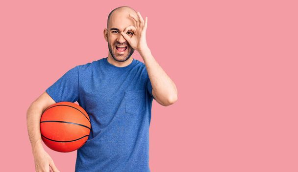 Fiatal jóképű férfi kezében kosárlabda mosolyogva boldog csinál oké jel kéz a szemen néz keresztül ujjait  - Fotó, kép