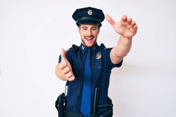 Молодой кавказский мужчина в полицейской форме смотрит на камеру, улыбаясь с распростертыми объятиями. веселое выражение, охватывающее счастье.  - Фото, изображение