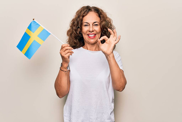 Idade Média bela turista segurando bandeira sueca sobre fundo branco isolado fazendo sinal ok com os dedos, sorrindo simpática gesto excelente símbolo
 - Foto, Imagem