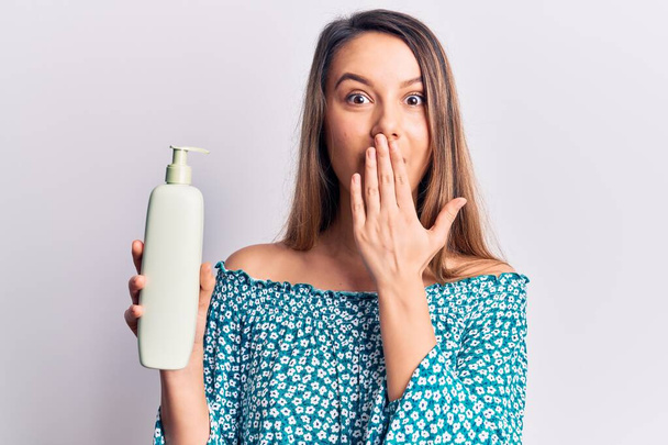 Young beautiful girl holding bottle of cream lotion covering mouth with hand, shocked and afraid for mistake. surprised expression  - Foto, Imagem