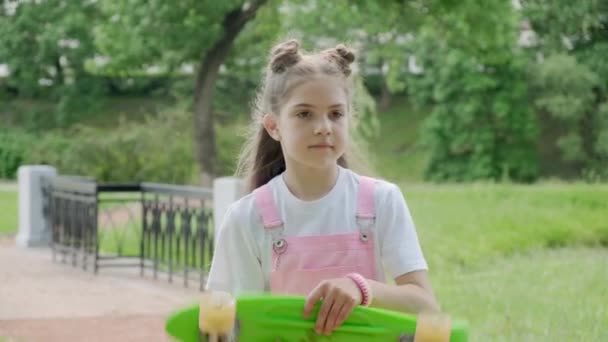 A beautiful young girl walks through the park and holds a scooter in her hand. - Video, Çekim