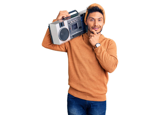 Handsome latin american young man holding boombox, listening to music looking confident at the camera smiling with crossed arms and hand raised on chin. thinking positive.  - Foto, Bild