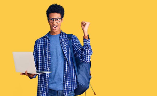 Νεαρός Αφροαμερικάνος που κρατάει φοιτητικό σακίδιο χρησιμοποιώντας φορητό υπολογιστή ουρλιάζοντας περήφανος, γιορτάζοντας τη νίκη και την επιτυχία πολύ ενθουσιασμένος με σηκωμένα χέρια  - Φωτογραφία, εικόνα