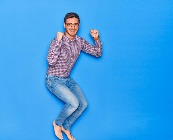 Young handsome hispanic businessman wearing glasses smiling happy. Jumping with smile on face celebrating with fists up over isolated blue background - Fotografie, Obrázek