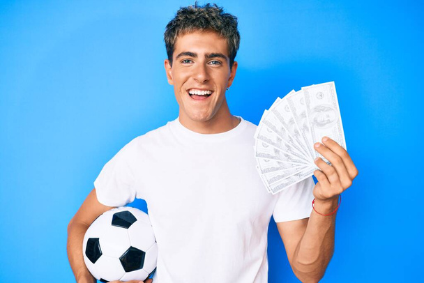 サッカーボールとドルの紙幣を持っている若いハンサムな男は笑顔で大声で笑います。.  - 写真・画像