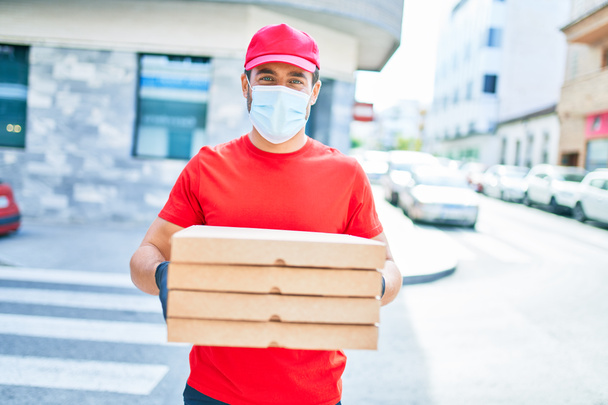 Young delivery man wearing uniform and coronavirus protection medical mask. Holding deliver pizza boxes at town street. - Photo, Image