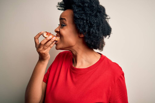 若い美しいアフリカ系アメリカ人のアフロ女性が笑顔で自信を持っています。白い背景の上にイタリアのピザのおいしいスライスを食べる顔に笑顔で立って - 写真・画像
