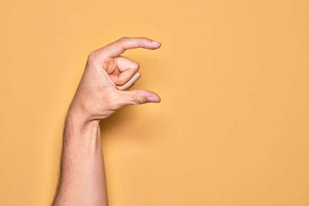 Hand of caucasian young man showing fingers over isolated yellow background picking and taking invisible thing, holding object with fingers showing space - Foto, Imagen
