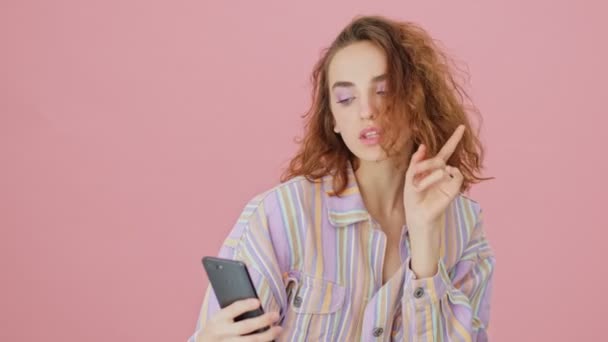 A close-up view of a cheerful young woman is taking selfie photos using her mobile phone standing isolated over a pink background in studio - Video